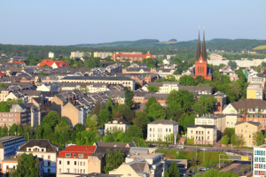 Stadtansicht mit Gebäuden, Kirche, Grünflächen und umliegender Landschaft.